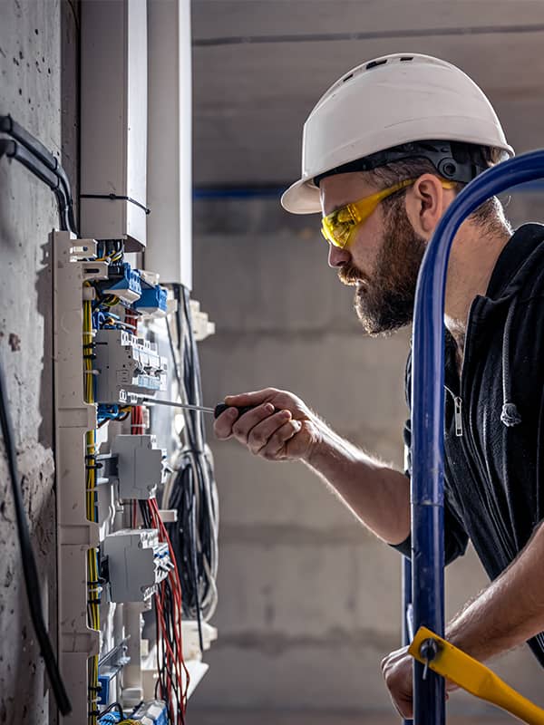 A male electrician works in a switchboard with a tool.