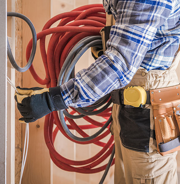 A construction worker in a plaid shirt and gloves holds electrical wires, with coiled hoses draped over their shoulder and a tool belt on their waist.