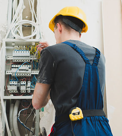 An electrician in a yellow hard hat works on a circuit breaker box, using tools and adjusting wires in a partially finished room.