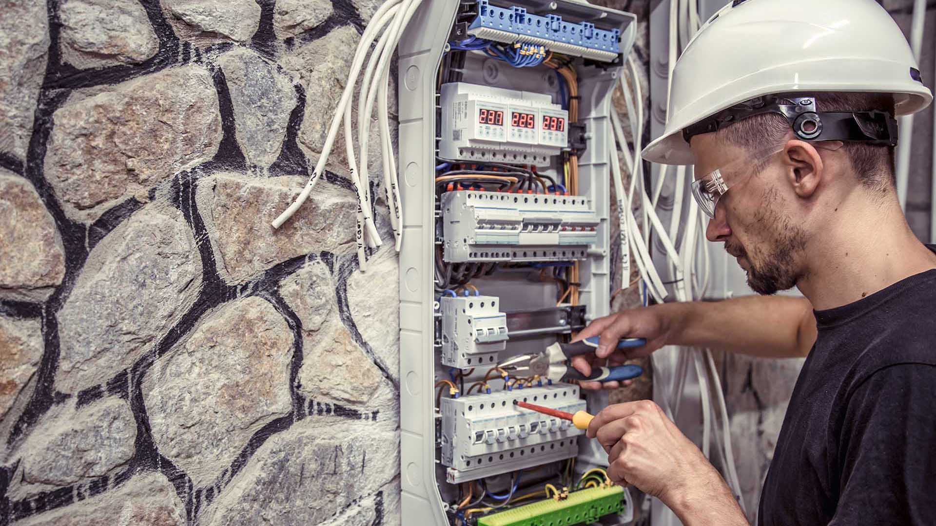 A technician in a hard hat works on an electrical panel surrounded by exposed wiring and a stone wall. Tools are in hand.