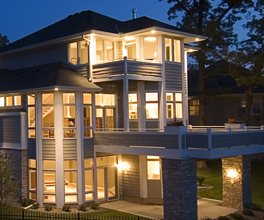 Modern two-story house illuminated at night, featuring large windows, a stone foundation, and a spacious balcony.