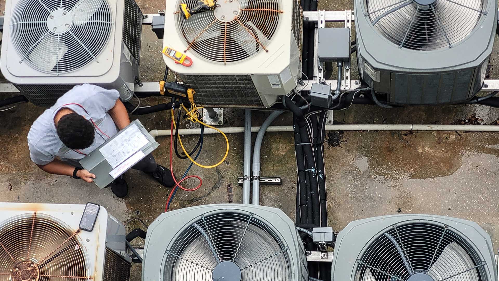 An HVAC technician inspects air conditioning units, holding a document, with multicolored wires and a smartphone nearby.