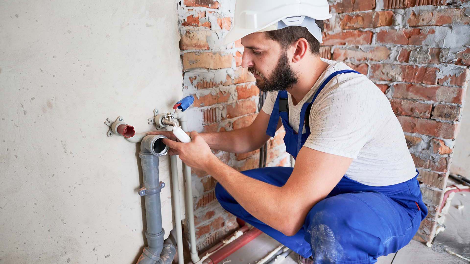 A worker in blue overalls and a safety helmet is crouched, connecting plumbing pipes against a brick wall in a construction setting.